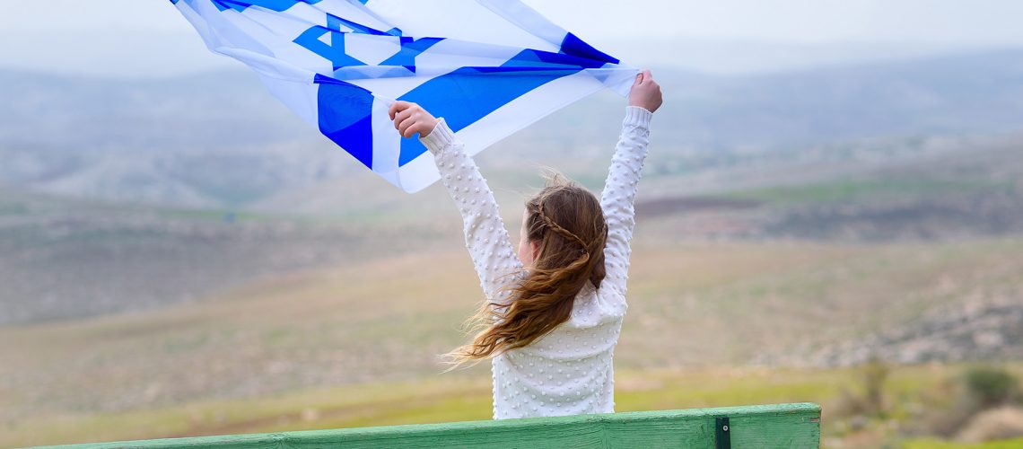 Beautiful young jewish girl sitting holding Israel flag in the wind and enjoying great view landscape on the sky, field and mountains.Patriotic holiday. Independence day Israel - Yom Ha'atzmaut .